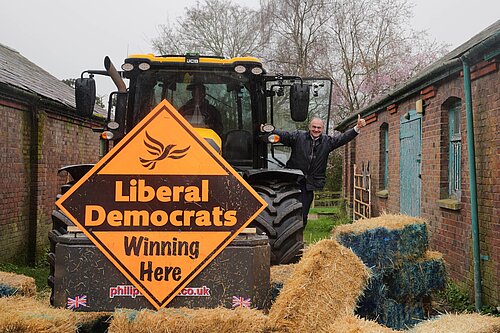 Ed Davey waves from tractor with Lib Dem sign, having smashed blue wall of bales